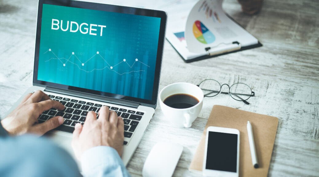 man working on laptop, word "budget" on screen. Coffee and smartphone next to laptop