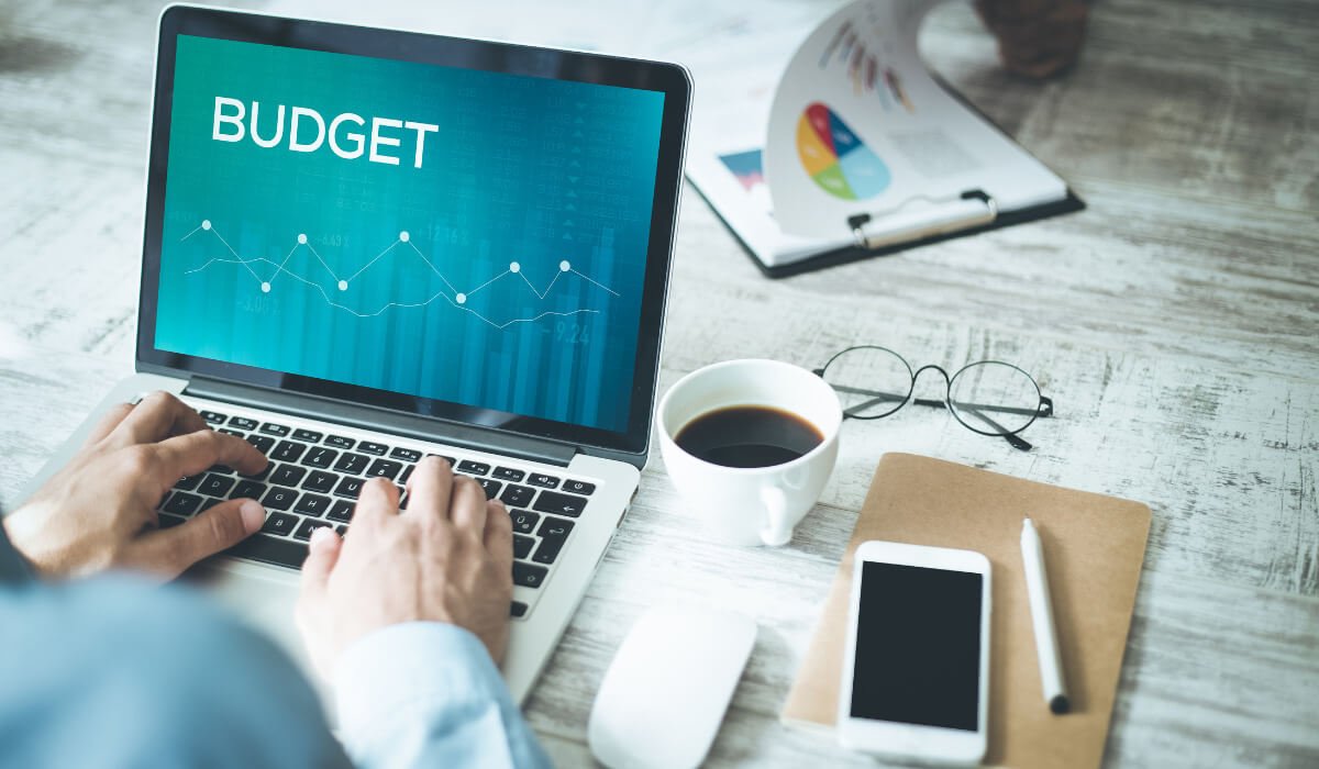 man working on laptop, word "budget" on screen. Coffee and smartphone next to laptop